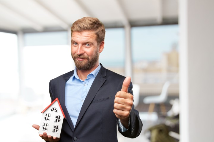 blond property manager in a suit showing thumbs up and holding a model house