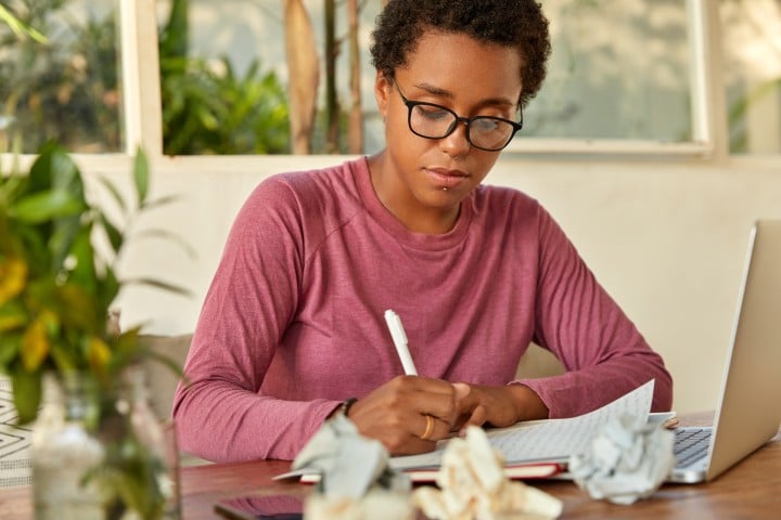 woman writing on sheet of paper with computer
